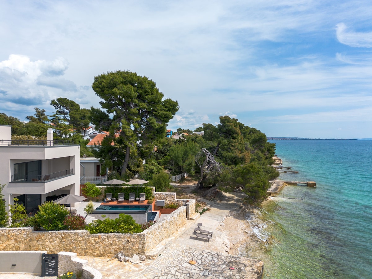 A view of the coastline highlights the proximity of the villa to the clear blue sea. Pebbly beaches can be seen alongside lush greenery and tall trees. The villa's outdoor space features a swimming pool area and sun loungers, enhancing the serene coastal environment.