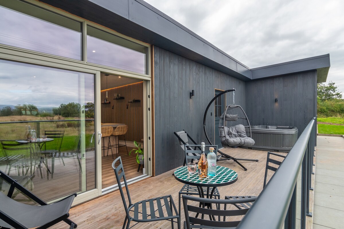 A spacious outdoor deck features a cozy seating area with a table and chairs, accompanied by a hanging chair. A hot tub is visible nearby, surrounded by greenery and expansive views of the countryside. Large glass doors lead inside, enhancing the outdoor connection.