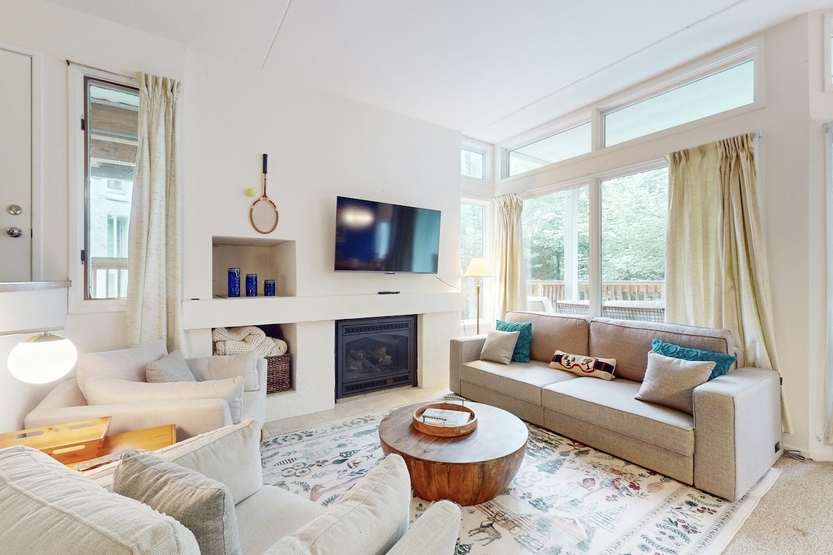 A bright living area features large windows that invite natural light, with a cozy seating arrangement including a gray sofa and armchairs. A wooden coffee table sits in the center, while a wall-mounted television and a gas fireplace enhance the inviting atmosphere.