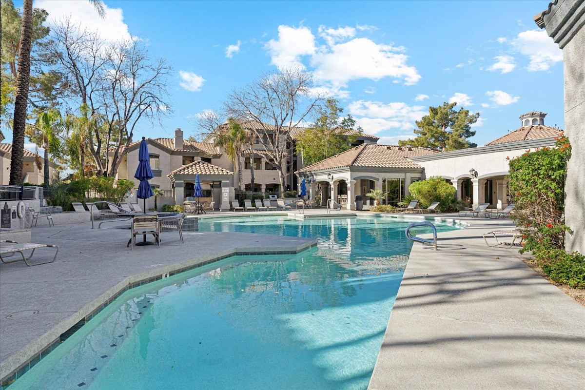 A large heated pool is surrounded by lounge chairs and palm trees, with several umbrellas offering shade. The serene environment is complemented by neatly maintained landscaping and a view of the nearby clubhouse, creating a relaxed atmosphere for guests.