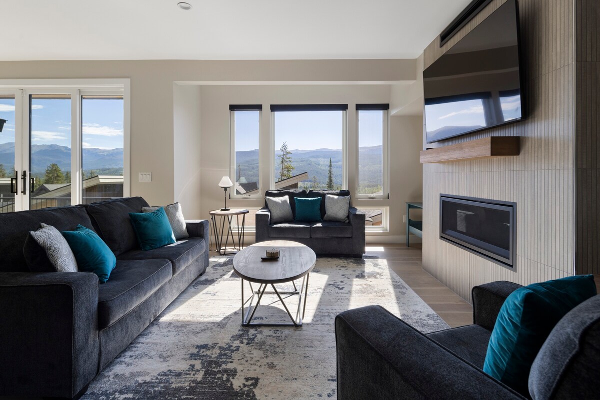 A modern living area features large windows with mountain views, allowing natural light to fill the space. Comfortable gray sofas are arranged around a circular coffee table, and a wall-mounted TV is positioned above a sleek fireplace. Decorative cushions add a touch of color.
