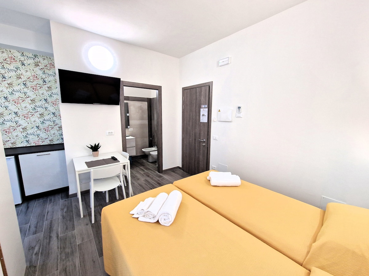 A bright double room features two single beds with yellow bedding arranged side by side. A wall-mounted TV is positioned above a small table and chairs. The private bathroom is visible through an open door, showcasing modern fixtures and a neutral color palette. A refrigerator is placed beside the table.