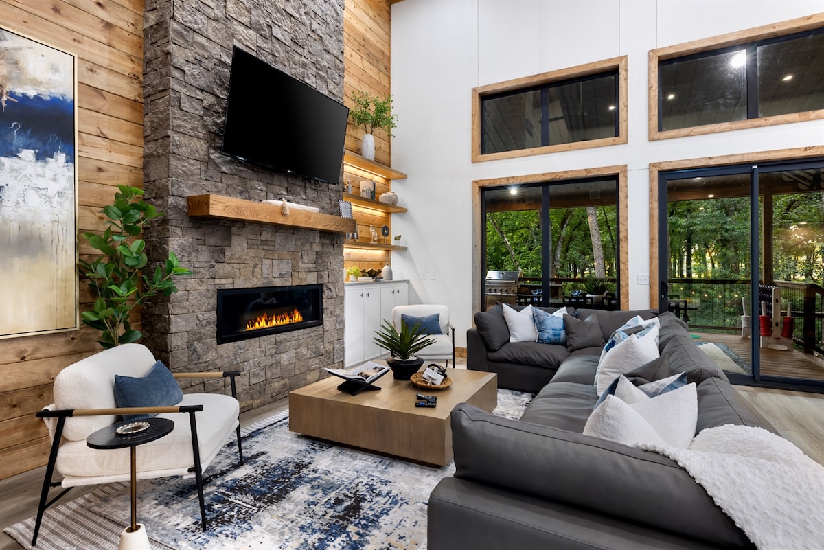 A modern living area features a decorative stone fireplace beneath a mounted TV. Comfortable gray and white sofas are arranged around a central coffee table, set on an area rug. Large windows provide views of the surrounding greenery, enhancing the spacious atmosphere.