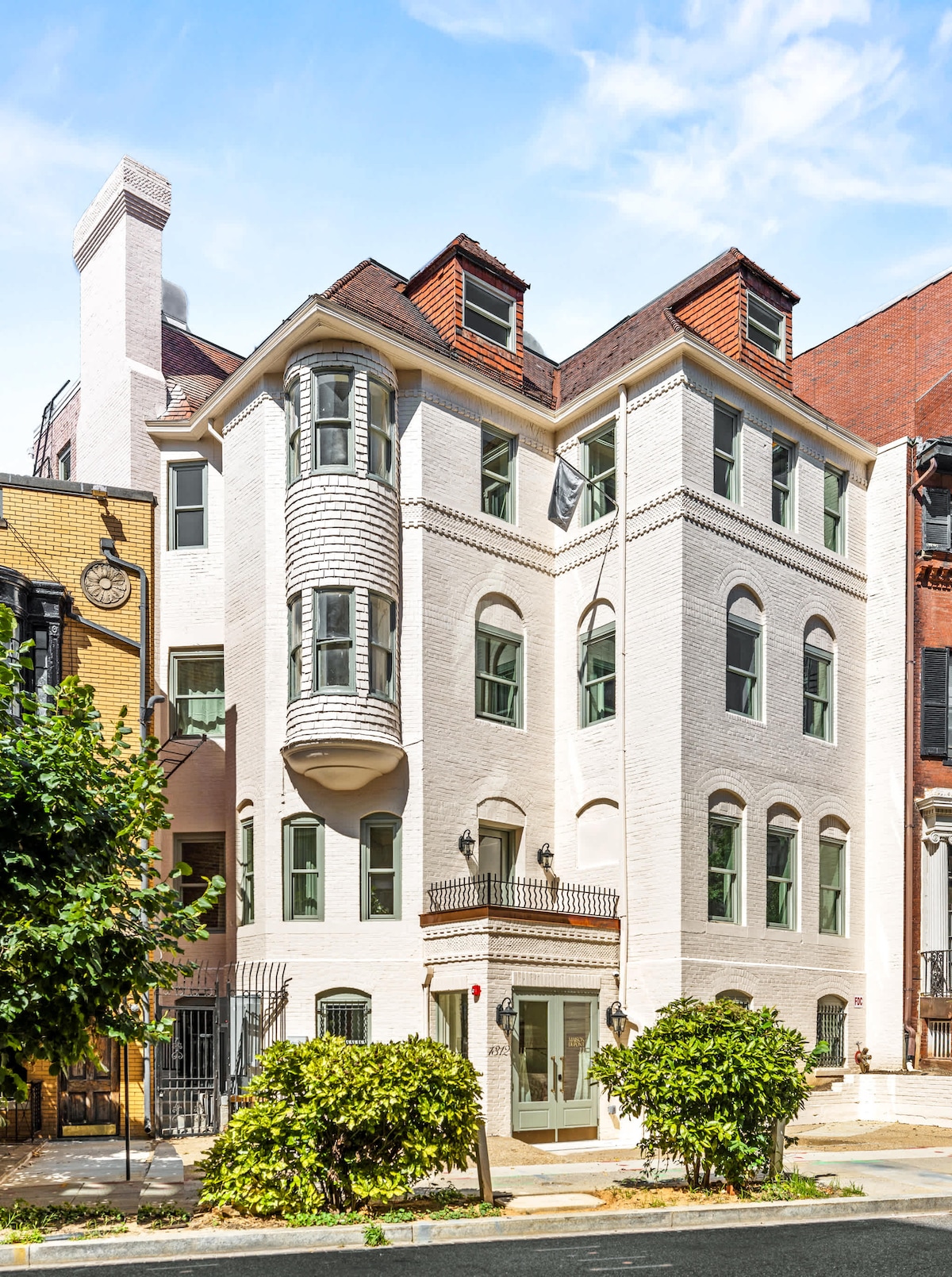 The exterior of the hotel displays a three-story building with brick walls painted in white. Large, symmetrical windows reflect the sunlight, while corner bay windows enhance the design. Framed by lush greenery, the entrance features a decorative balcony supported by elegant columns.