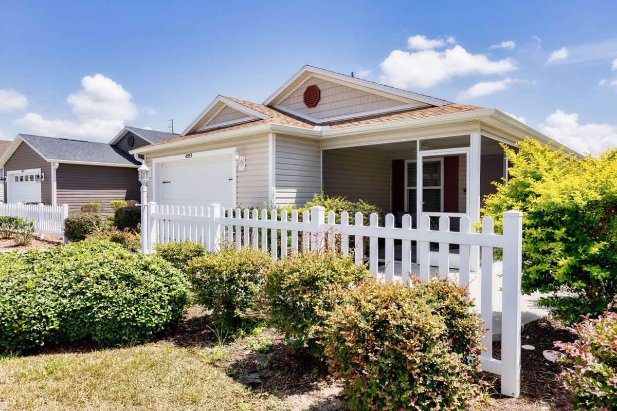 A welcoming exterior view of the villa is framed by a white picket fence and vibrant landscaping. The home features a single-car garage, a covered entryway, and large windows that allow natural light to illuminate the interior. The clear blue sky enhances the cheerful atmosphere.