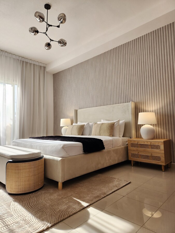 A light-filled bedroom features a spacious king-sized bed adorned with decorative pillows. Two bedside lamps provide soft illumination. A stylish bench rests at the foot of the bed, while large windows with sheer curtains invite natural light, highlighting the textured wall behind the headboard.