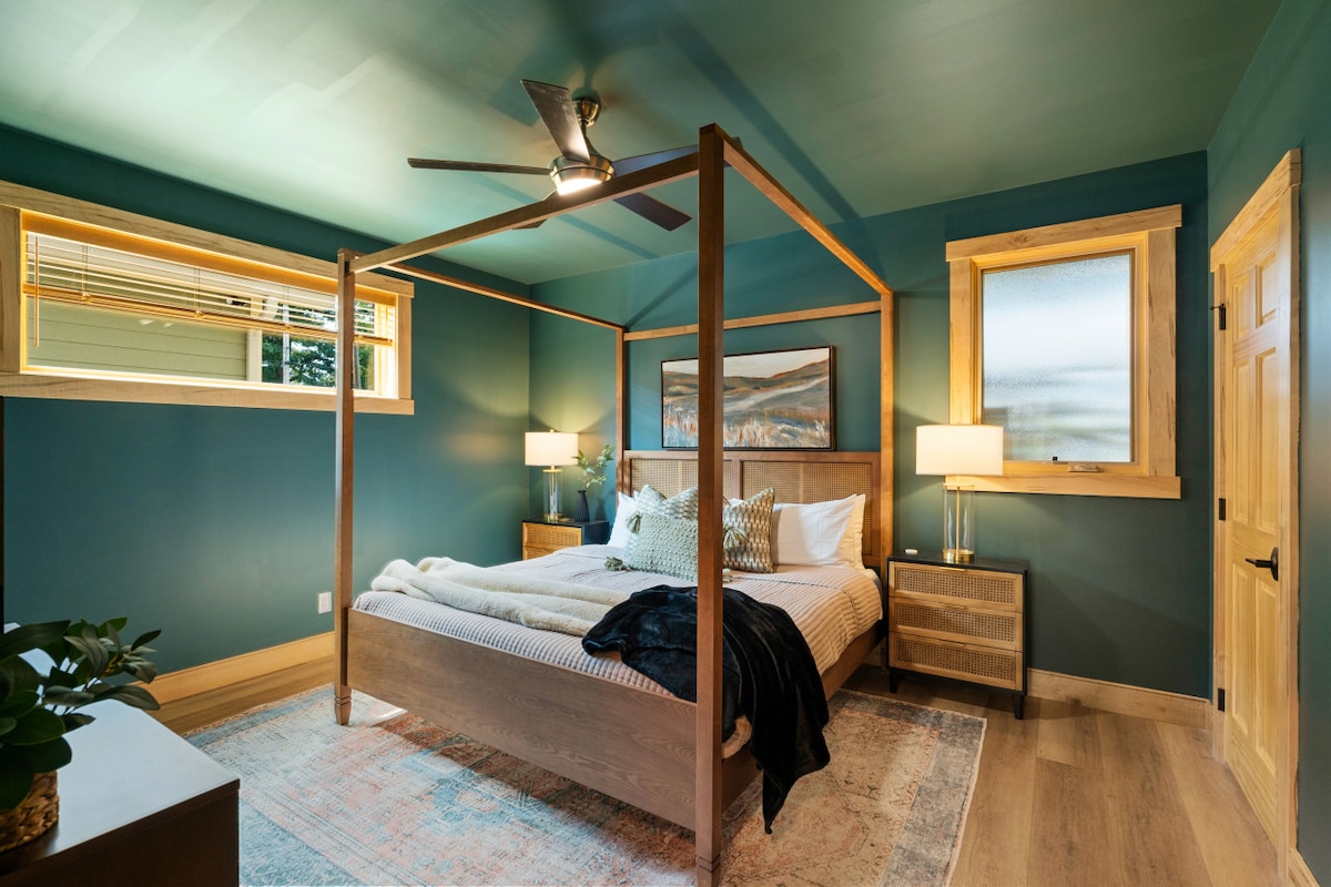 A cozy bedroom features a four-poster bed with a light-colored quilt. Soft lighting is provided by two bedside lamps on matching nightstands. The walls are painted a soothing green, and natural light enters through two windows. A textured rug adds warmth to the space.