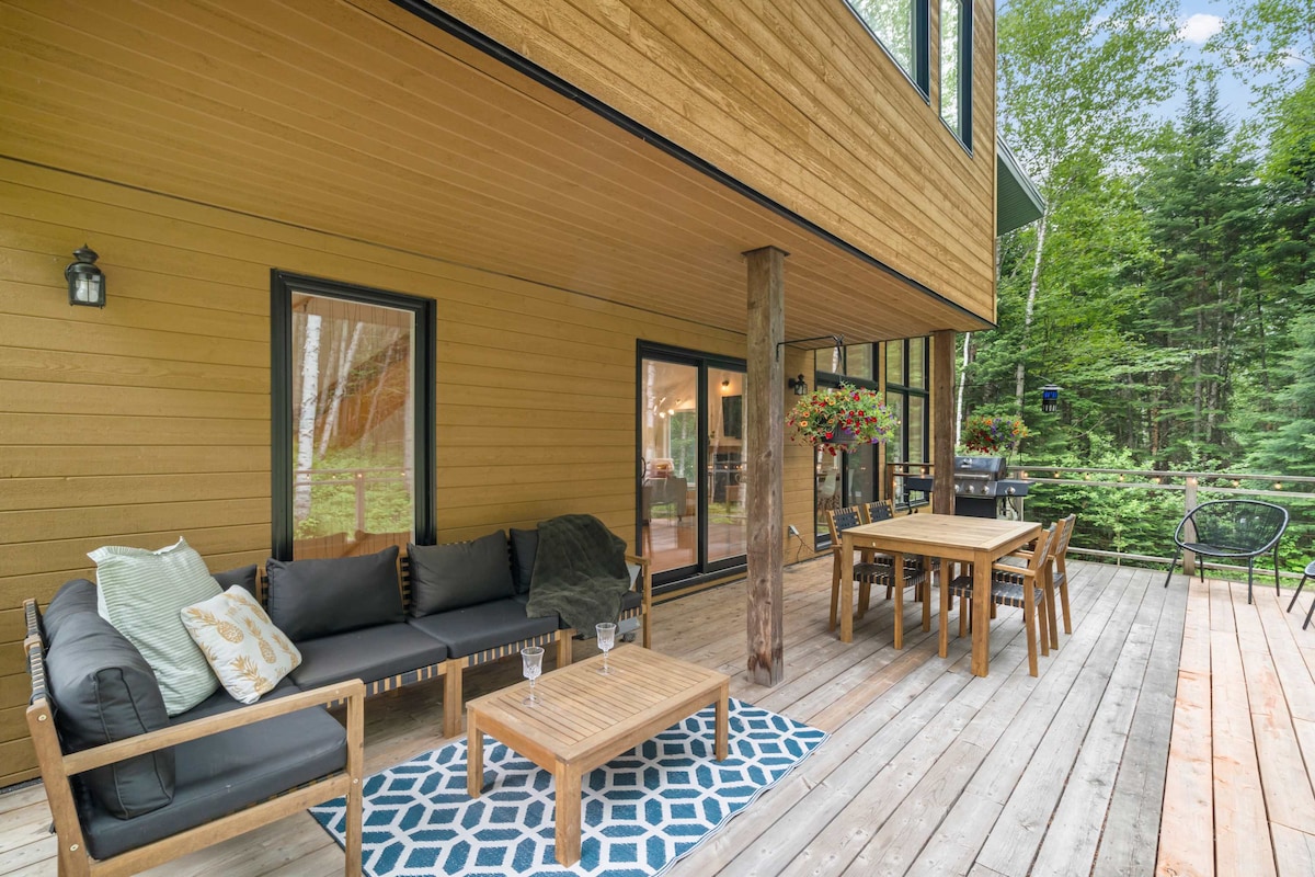 A spacious outdoor patio is presented, featuring a comfortable seating area with a dark sofa and a geometric rug. A wooden dining table with chairs complements the natural surroundings, while large glass doors provide views of the interior. Lush greenery creates a serene backdrop.