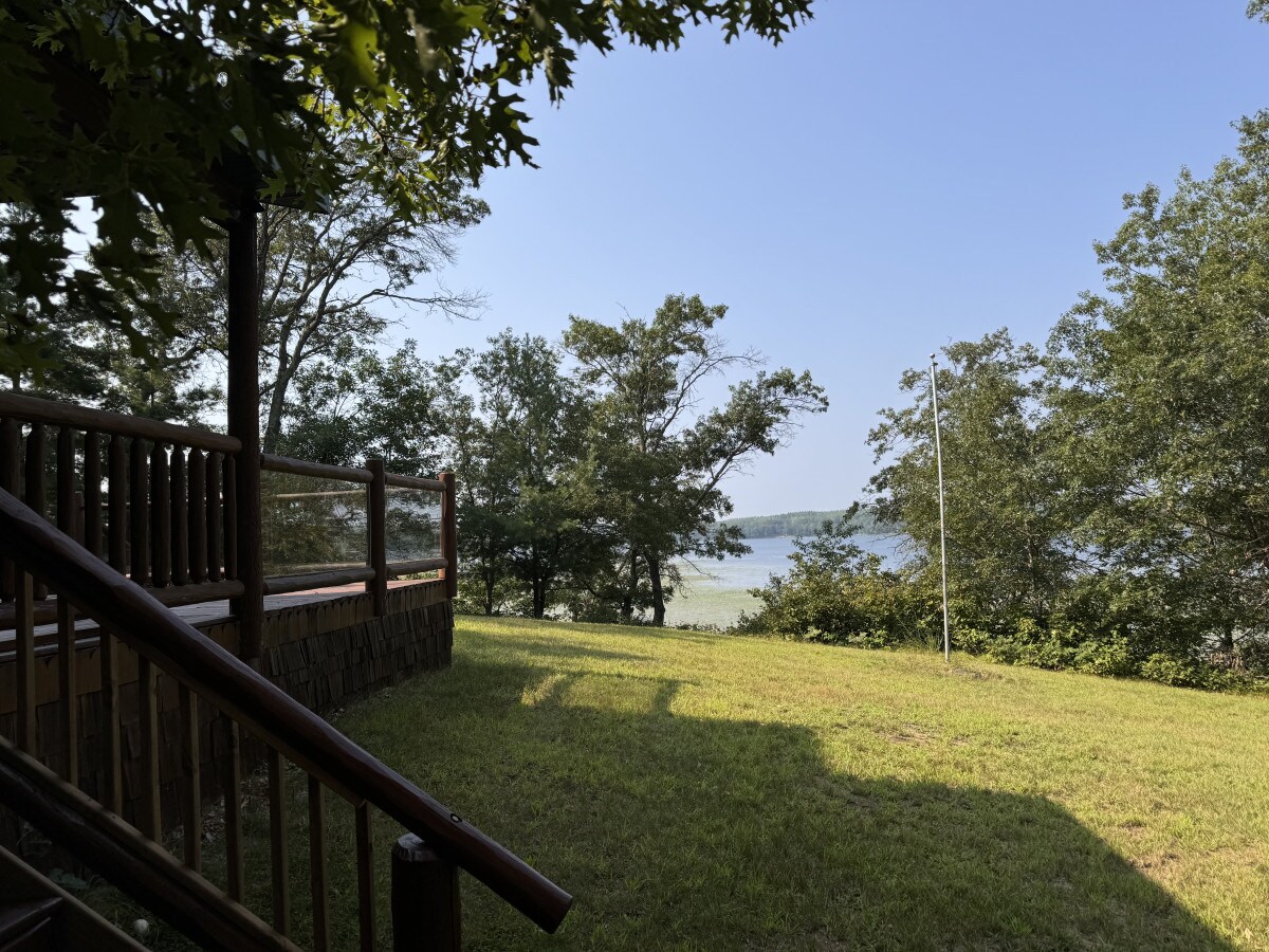 A view of lush green grass leading to serene waters, framed by trees. A wooden deck is visible in the foreground, offering a relaxing vantage point to enjoy the natural surroundings.