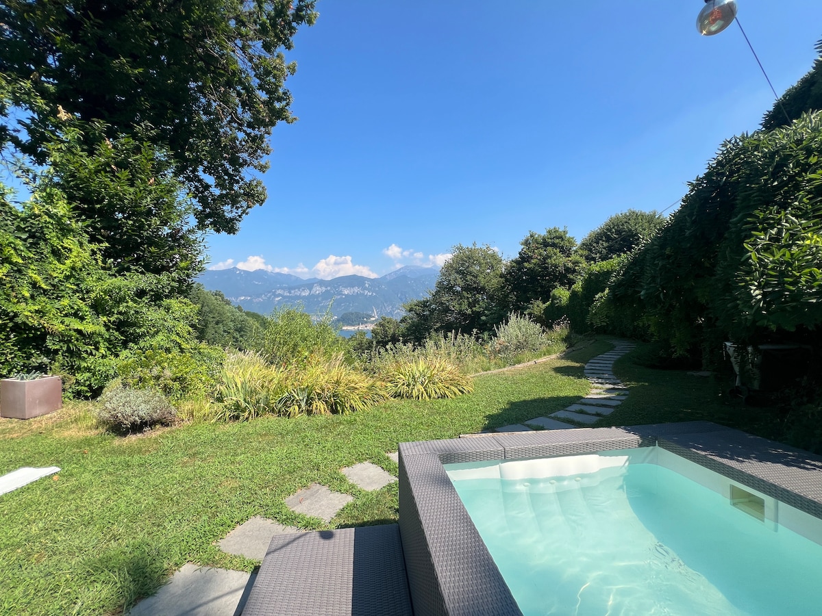 An outdoor space featuring a heated jacuzzi surrounded by a lush green lawn and neatly arranged stone pathways. The view extends to Lake Como and distant mountains, under a clear blue sky, creating a serene atmosphere that invites relaxation.