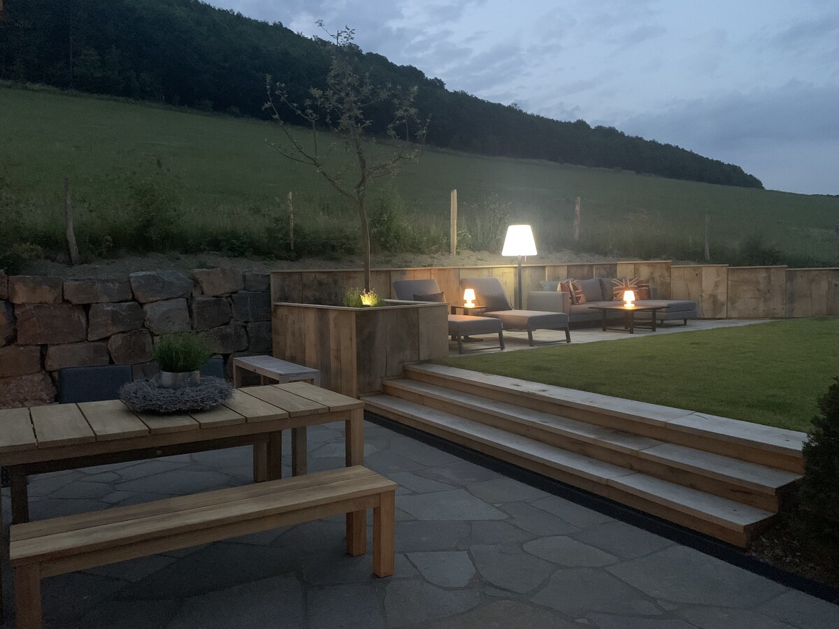 An outdoor seating area is illuminated softly in the evening light. Comfortable furniture is arranged on a stone patio, with a coffee table centerpiece. Steps lead to a lush green lawn and a natural stone wall serves as a background, enhanced by gentle lighting.