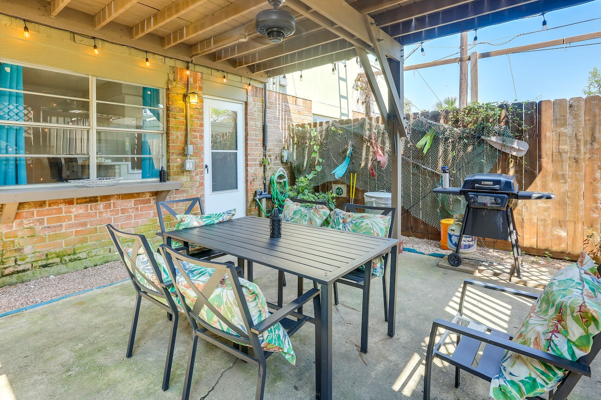 An outdoor dining area features a large table surrounded by six seats with tropical-patterned cushions. A gas grill is positioned nearby. String lights hang overhead, enhancing the inviting ambiance. The area is enclosed by wooden fencing and greenery, providing a private setting.