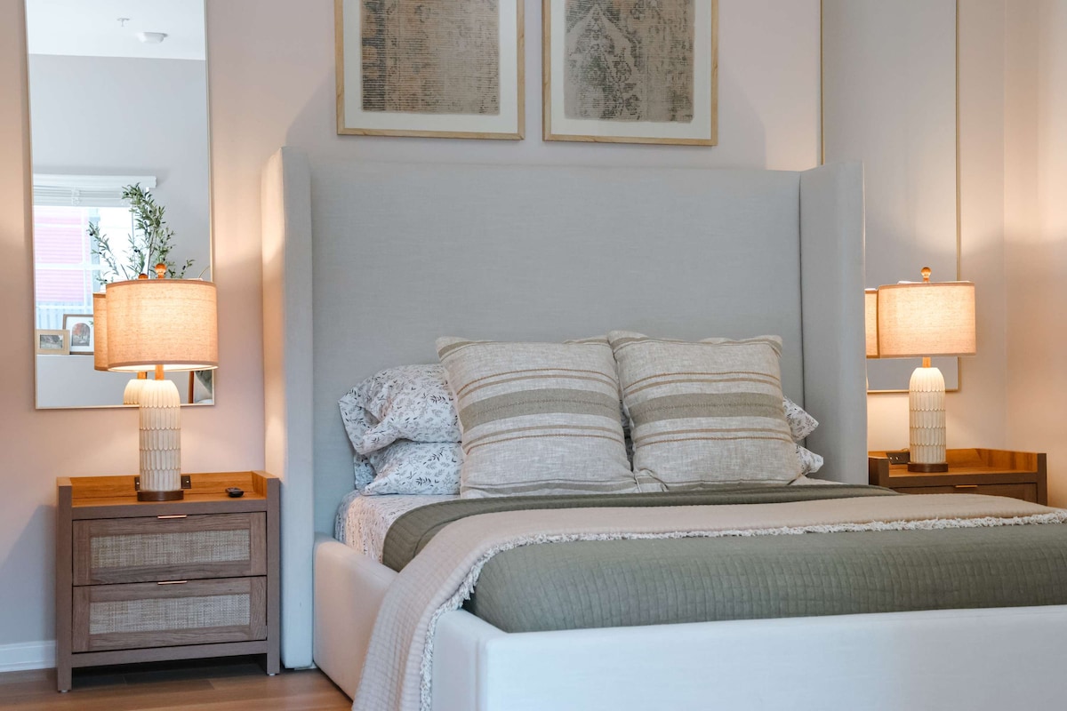 A stylish bedroom features a spacious bed with layered bedding and decorative pillows. Warm-toned lamps sit on each side of modern bedside tables. Artwork adorns the wall above, and a mirror reflects natural light from the room.