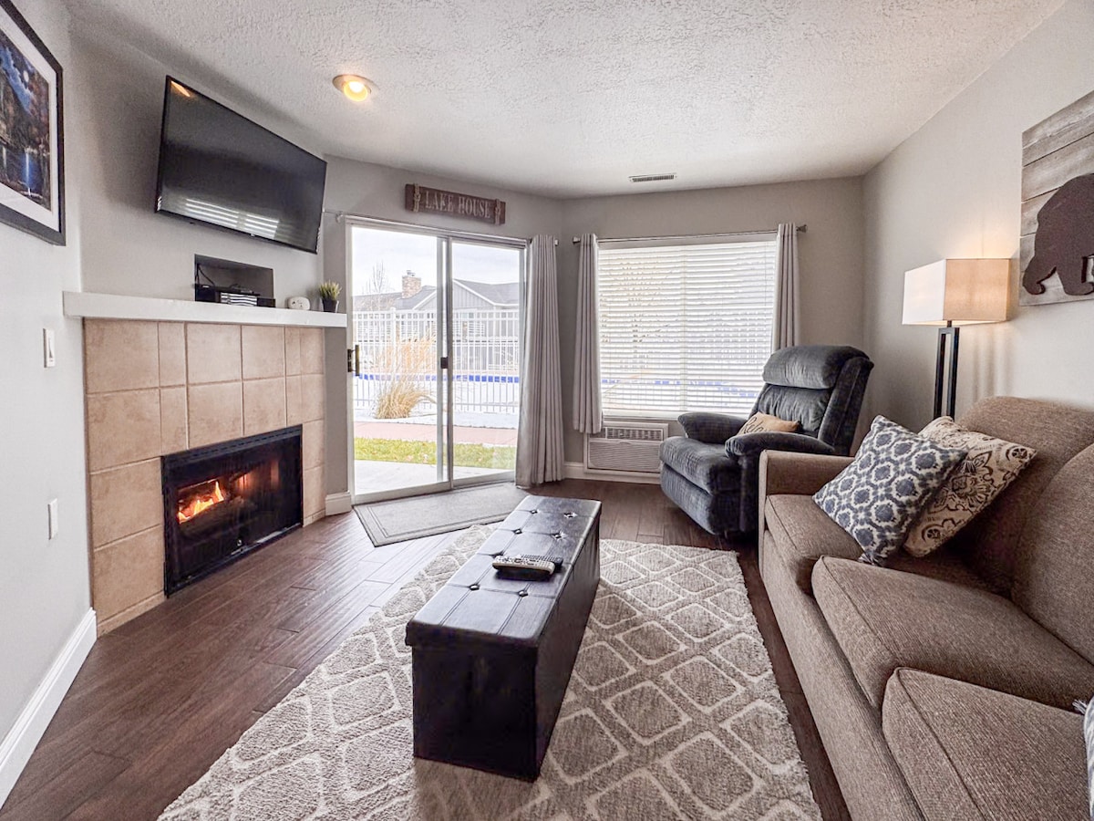 A cozy living area is set with a comfortable sofa and chair. A flat-screen TV is mounted above a decorative fireplace. Natural light enters through a large window with sheer curtains, offering a view of the pool area. A coffee table is positioned centrally.