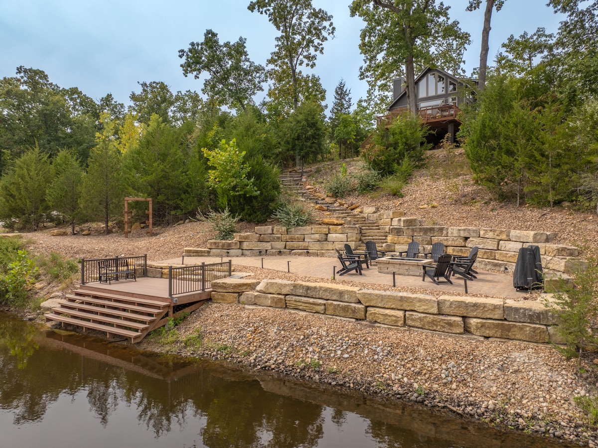 The outdoor space features a tiered patio area set along a serene lakeshore, furnished with several black lounge chairs and a dining set. A stone walkway leads up to the house, surrounded by lush greenery and trees. A composite dock is positioned for easy lake access.