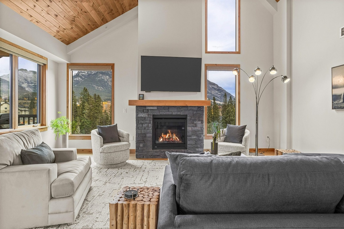 A spacious living area is highlighted by tall windows offering panoramic views of the surrounding nature. A gas fireplace with a stone surround serves as the focal point, while comfortable seating options, including two cozy armchairs and a large sectional sofa, invite relaxation.
