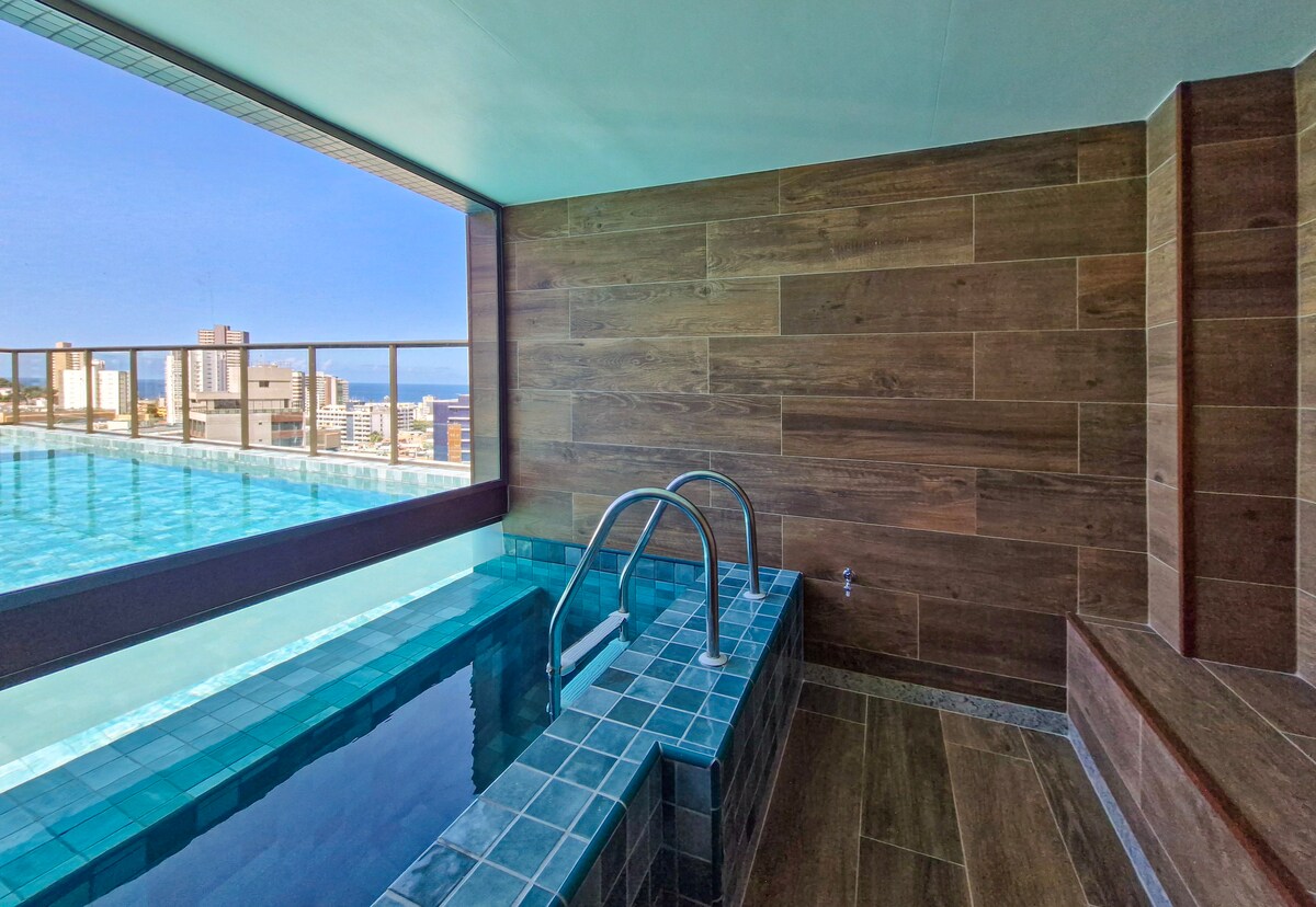 An indoor pool area features large windows that provide a panoramic view of the city and ocean. The pool is framed by natural wood paneling, and steps lead into the water from a tiled sensation platform, creating a relaxed and inviting atmosphere.
