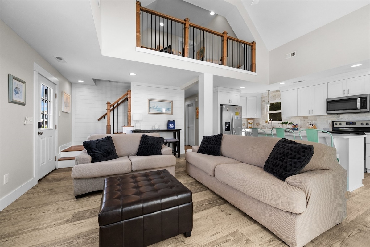 The open-concept living area features two sofas, a coffee table, and a staircase leading to the loft. Natural light floods the space through large windows, highlighting the modern kitchen equipped with updated appliances and a dining area with bar seating, creating an airy and inviting environment.