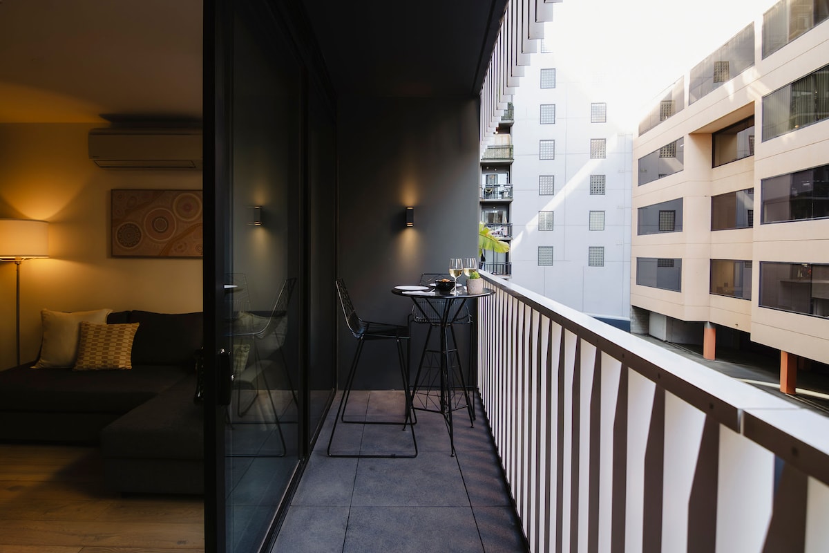 A modern balcony offers a glimpse of urban living with sleek furniture arranged for alfresco dining. A small table with high chairs is positioned against a glass railing, while soft ambient lighting enhances the contemporary space. Views of surrounding buildings create a vibrant city atmosphere.