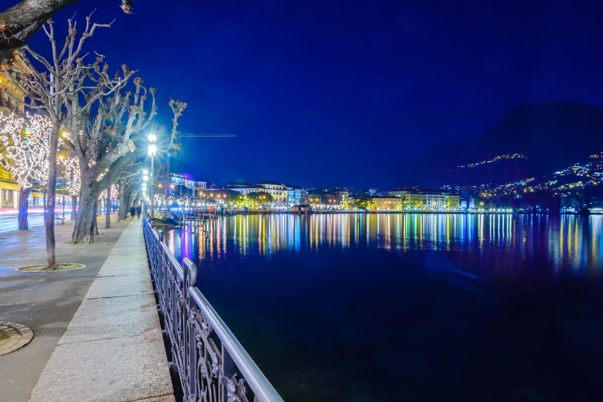 The lakeside promenade is shown during the evening, with lights reflecting off the calm water. Illuminated buildings line the water's edge, and trees with decorations provide a festive touch. The surrounding hills are subtly visible under the night sky.