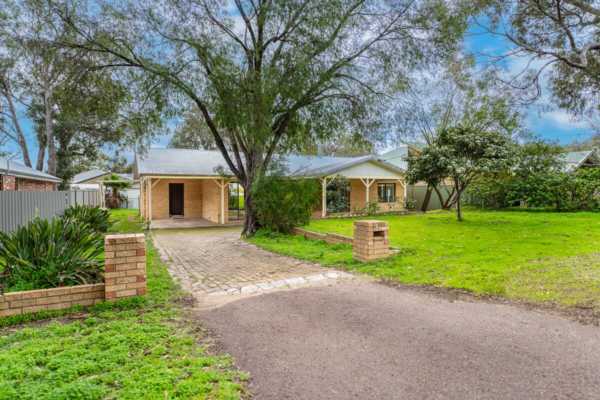 A well-maintained exterior of a single-story home is surrounded by green grass and mature trees. A paved pathway leads to the front porch, and the entryway is framed by a protective overhang.