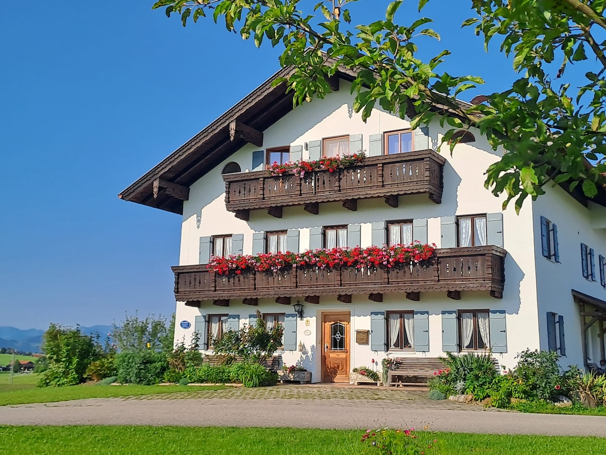 A charming rural building features multiple balconies adorned with vibrant red flowers. The facade presents a combination of traditional wooden elements and light-colored walls, set against a clear blue sky. Lush greenery surrounds the property, enhancing its serene countryside setting.