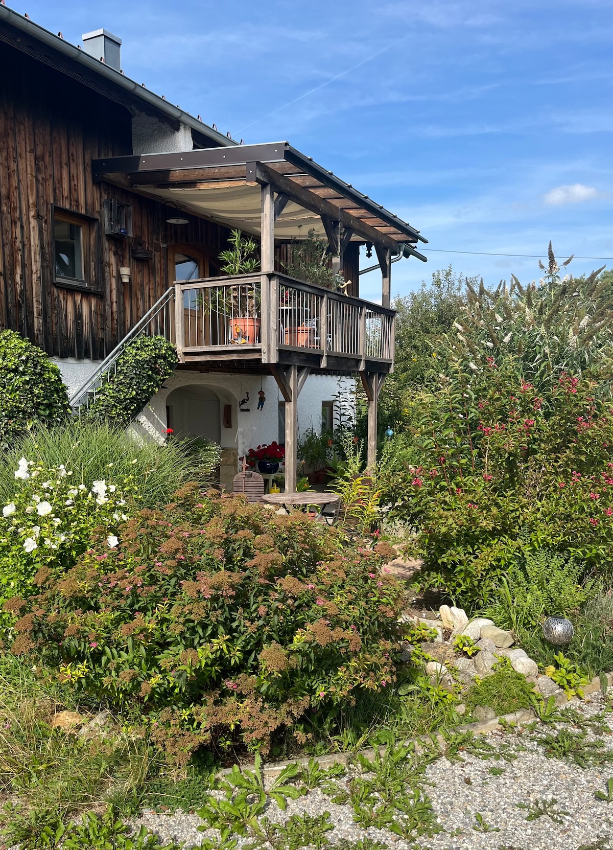 The image shows a wooden house with a covered balcony supported by a staircase. Surrounding the entrance are various plants, including white flowers and greenery. The pathway is lined with rocks, contributing to the natural ambiance of the outdoor space.