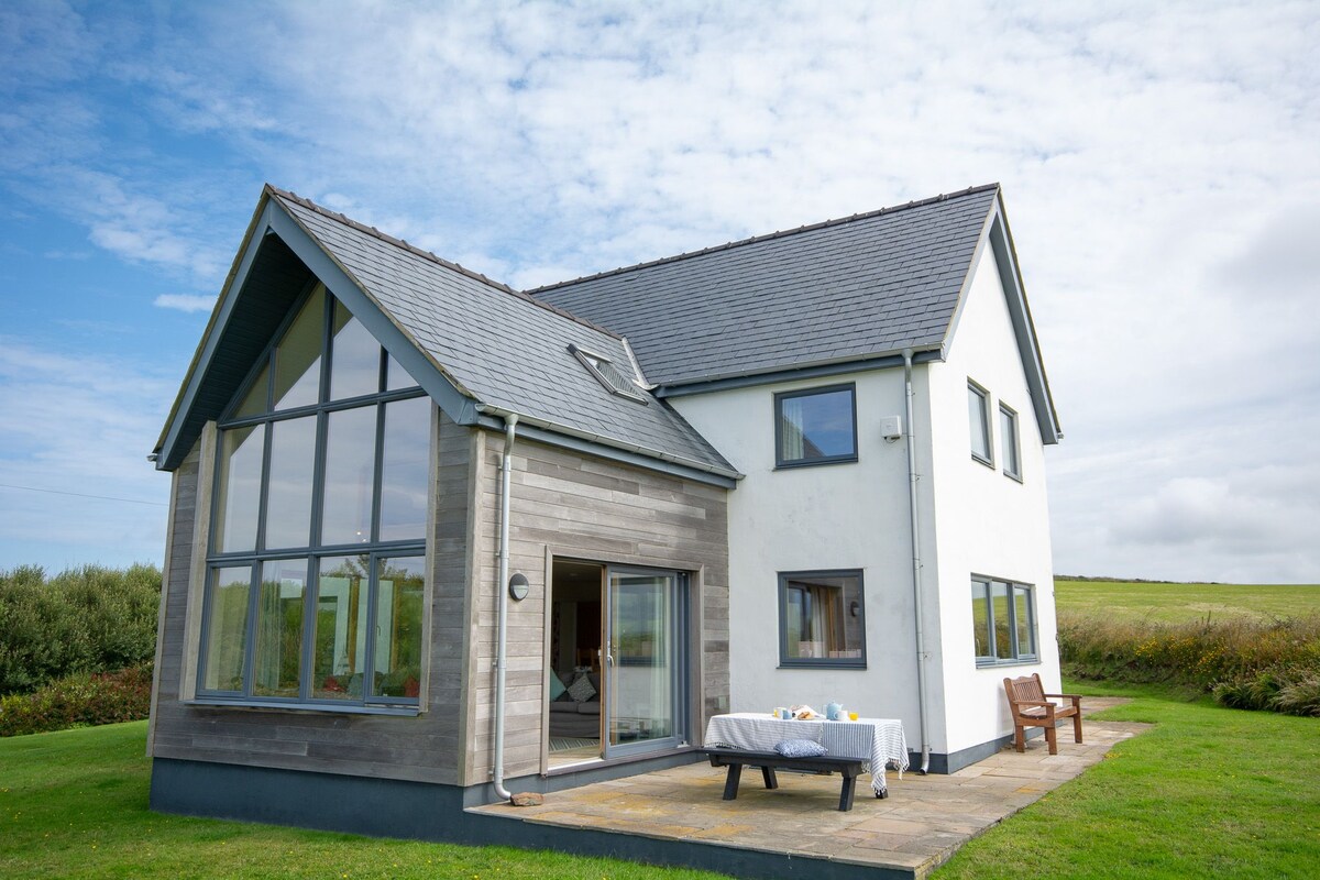 A modern two-story house with a combination of wood and white exterior stands against a bright sky. Large windows allow ample light into the space, while a stone patio area features a table set for meals. Lush greenery surrounds the property.