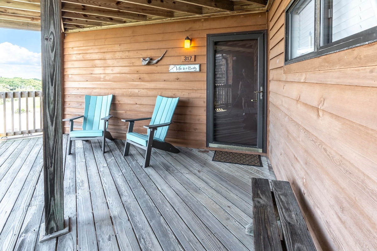 An inviting wooden deck features two light blue Adirondack chairs positioned for relaxation. A dark front door with a decorative light is visible, framed by a natural wood exterior. The flooring is made of weathered planks, creating a rustic atmosphere.