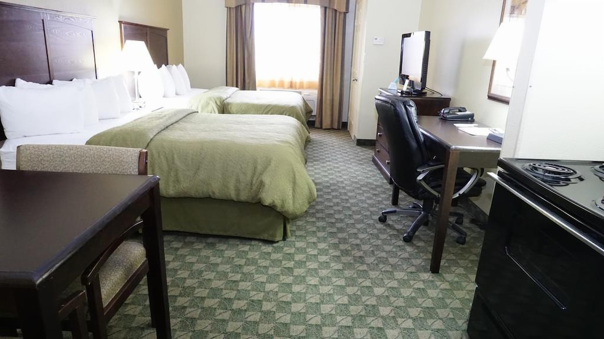 A hotel room features two Queen beds dressed in light green bedding, accompanied by a desk with an office chair and a small dining table. Natural light enters through the window, which is fitted with light-filtering curtains. A TV and microwave are also visible.