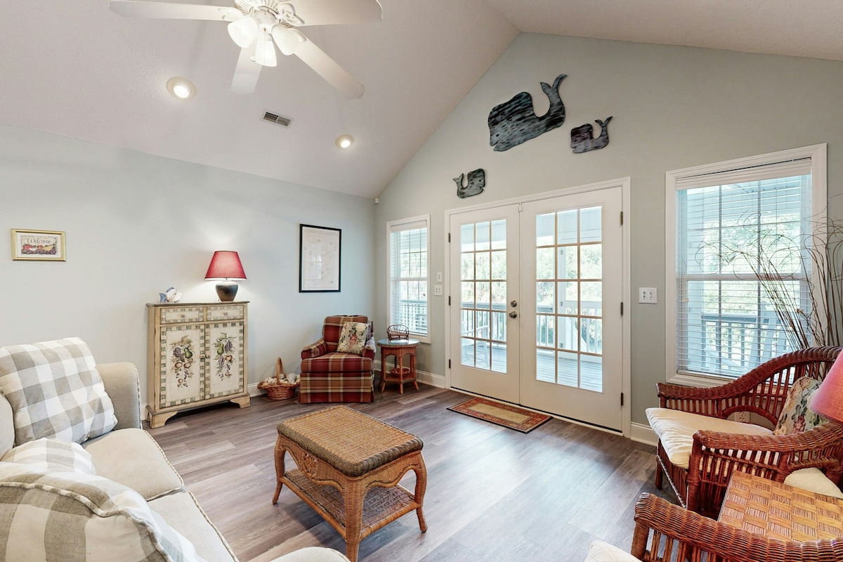 A bright living room features a high cathedral ceiling with ceiling fan. Comfortable seating options include a sofa and wicker chairs. A decorative cabinet adds character, while natural light flows in through glass doors that lead to the outside. A cozy area rug defines the space.