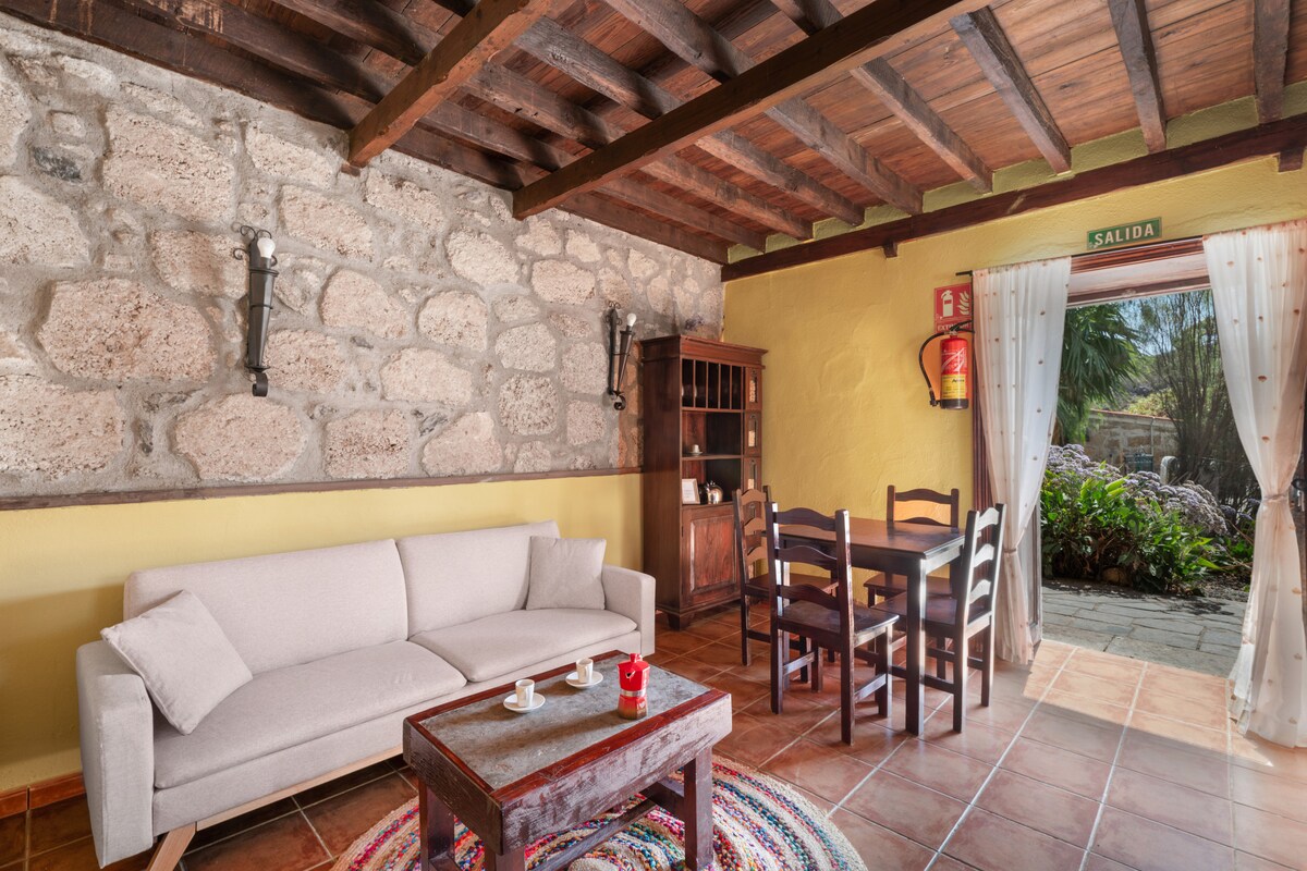 A living area features a light-colored sofa set against a textured stone wall. A wooden dining table with chairs is positioned nearby. Natural light enters from the open door, highlighting the room's neutral tones and traditional decor elements.