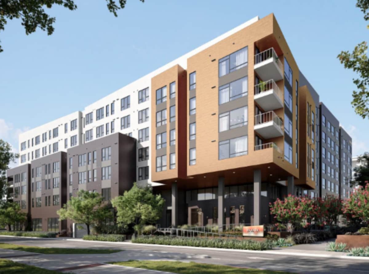 The modern building features a mix of light and dark exterior materials, with large windows and balconies on multiple floors. Lush green landscaping with shrubs and trees borders the entrance, promoting a welcoming environment near the street.