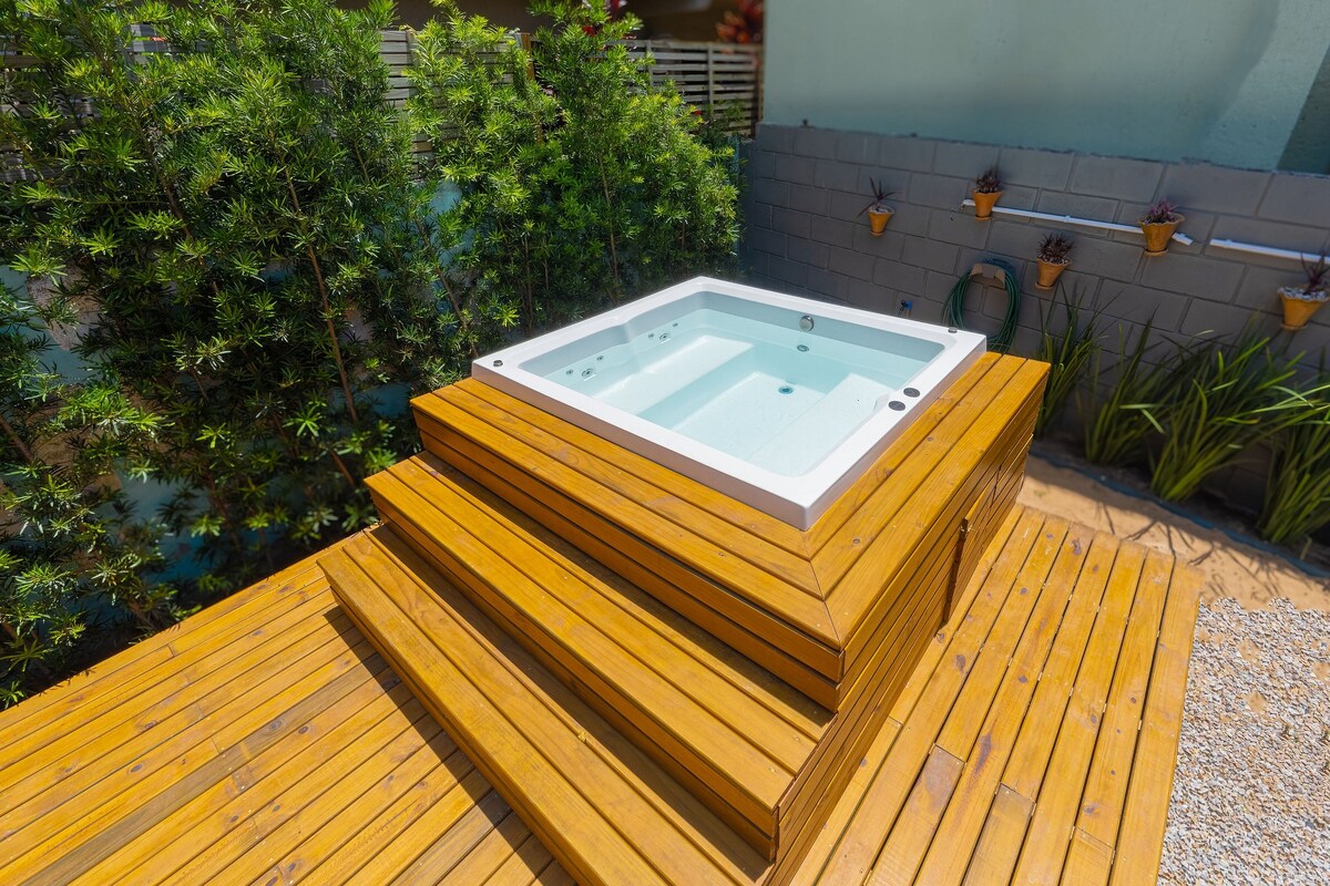 A hydromassage jacuzzi is positioned on wooden decking, surrounded by lush greenery. The jacuzzi features a clean, inviting surface, with steps leading up to it, enhancing accessibility. Small wall-mounted planters can be seen in the background, adding to the tranquil outdoor setting.