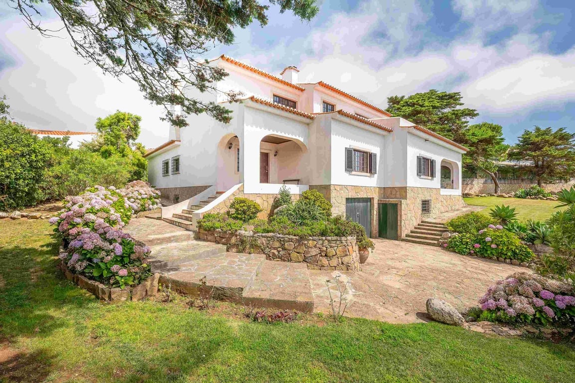 A spacious three-story house is featured, surrounded by a well-maintained garden filled with colorful flowers. Stairs lead toward an inviting entrance, while lush greenery frames the property. The light-colored exterior contrasts beautifully with the blue sky and scattered clouds above.