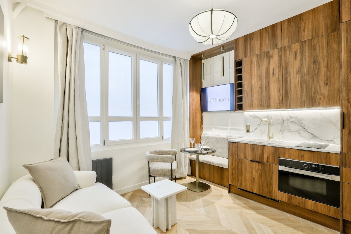 A modern living area features large windows allowing natural light to filter in. A beige sofa is positioned next to a small table with two elegant chairs. The cabinetry showcases a sleek design, highlighted by a marble countertop and wood finish, enhancing the overall aesthetic.