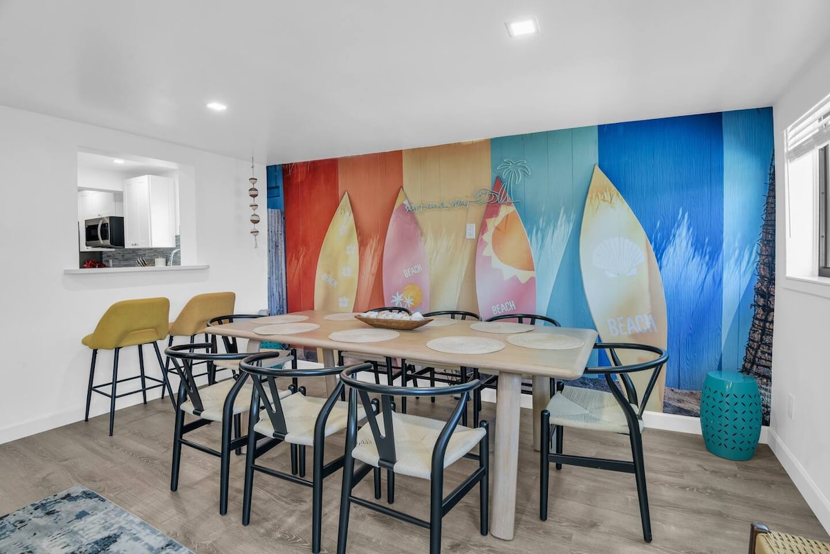 A dining area features a spacious wooden table surrounded by six upholstered chairs. A colorful beach-themed mural is displayed on the wall, depicting surfboards against a bright backdrop. Additional seating is provided at a bar counter with two stools in the background.