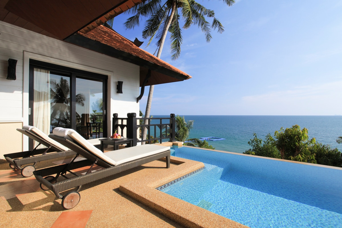 A private terrace features an inviting infinity pool that overlooks the Andaman Sea. Two sun loungers are positioned for relaxation, and palm trees frame the serene view, enhancing the connection with nature.