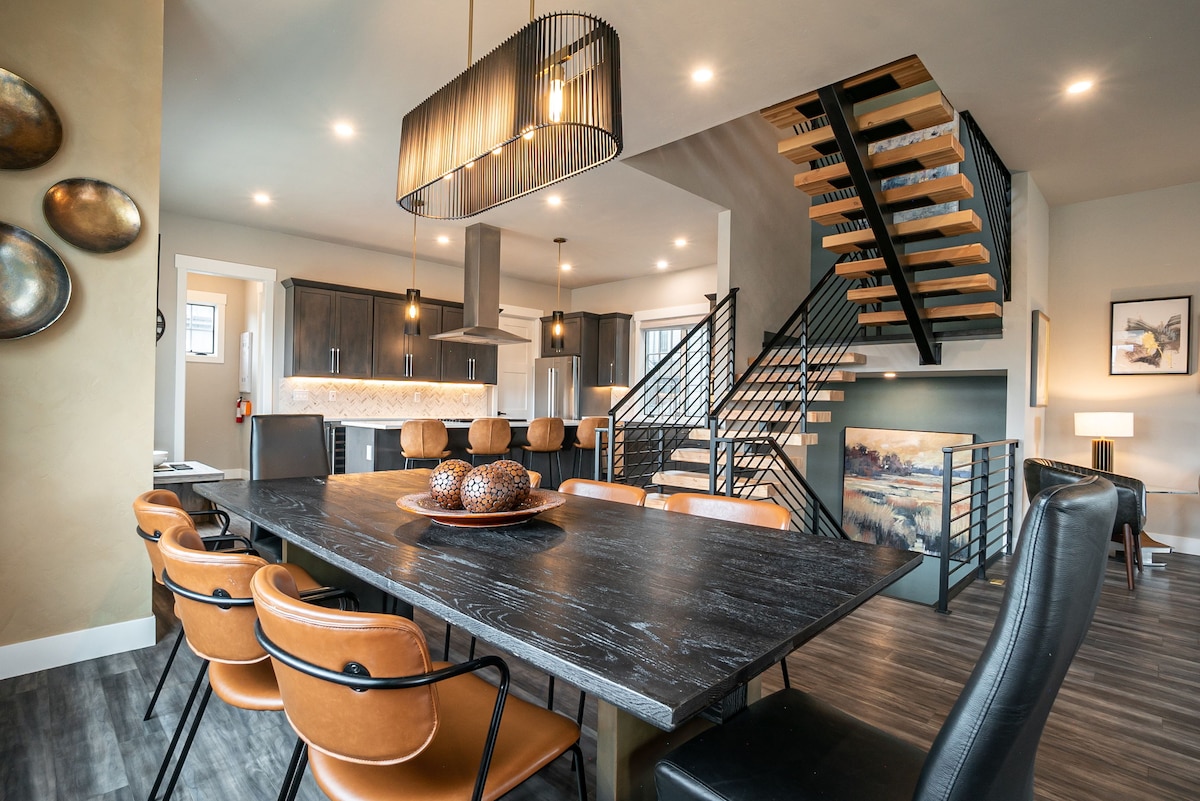 An open-concept dining area features a rustic table surrounded by leather chairs. A modern pendant light hangs overhead, illuminating the space. In the background, a sleek kitchen showcases dark cabinetry and a large island, while a stylish staircase leads to the upper level.