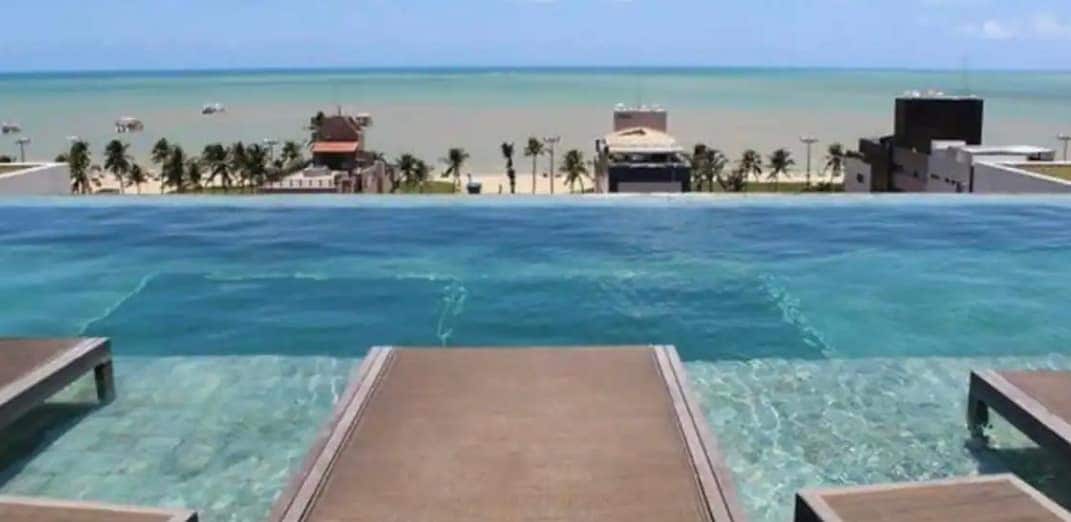 An infinity pool overlooks the beach, with clear blue waters merging seamlessly with the sky. Sun loungers are positioned along the edge, providing a space for relaxation. Palm trees sway gently in the background, framing the scenic coastal view.