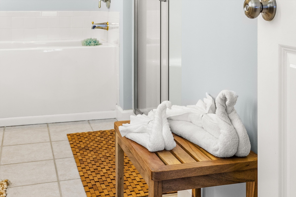 A neatly arranged wooden stool holds freshly folded white towels, shaped like swans. Light blue walls surround the space, accompanied by neutral tiled flooring. A spacious soaking tub and glass shower enclosure are visible in the background, enhancing the bathroom's tranquil aesthetic.