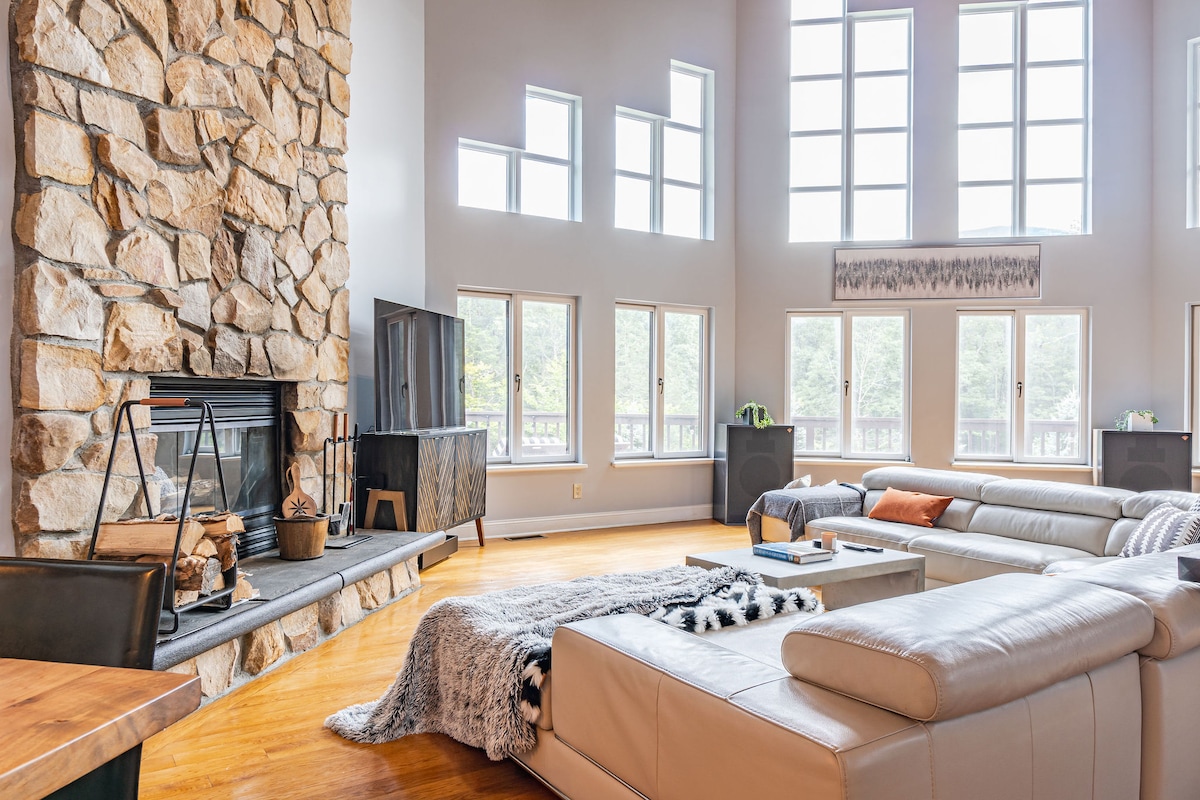 A spacious living room showcases a tall stone fireplace as its central feature. Natural light floods in through large windows, illuminating the comfortable seating arrangement. Stylish furniture, including cream-colored sofas and a rustic coffee table, is complemented by soft textures, creating an inviting space for relaxation.