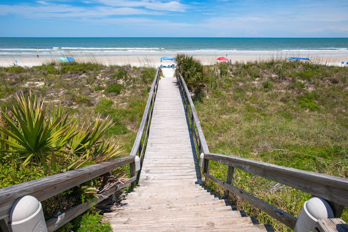 A wooden boardwalk leads towards the sandy beach, flanked by lush greenery on either side. The ocean is visible in the distance, with beach umbrellas set up along the shoreline. Soft clouds drift across a blue sky, creating a serene atmosphere.