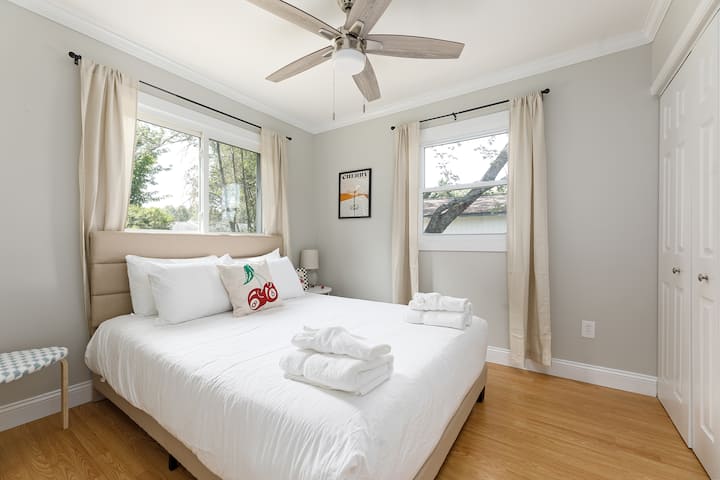 Chambre ensoleillée avec literie moelleuse, ventilateur de plafond et vue sur les feuilles à travers de grandes fenêtres.