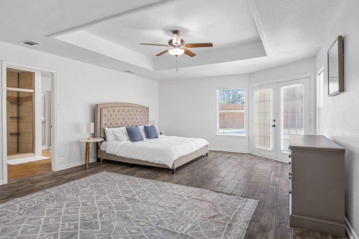 A spacious primary bedroom features a stylish bed with a tufted headboard and coordinating accent pillows. Natural light fills the room through large windows, while a ceiling fan provides ventilation. The en-suite bathroom is visible through an open door, and patio doors lead to the pool area.