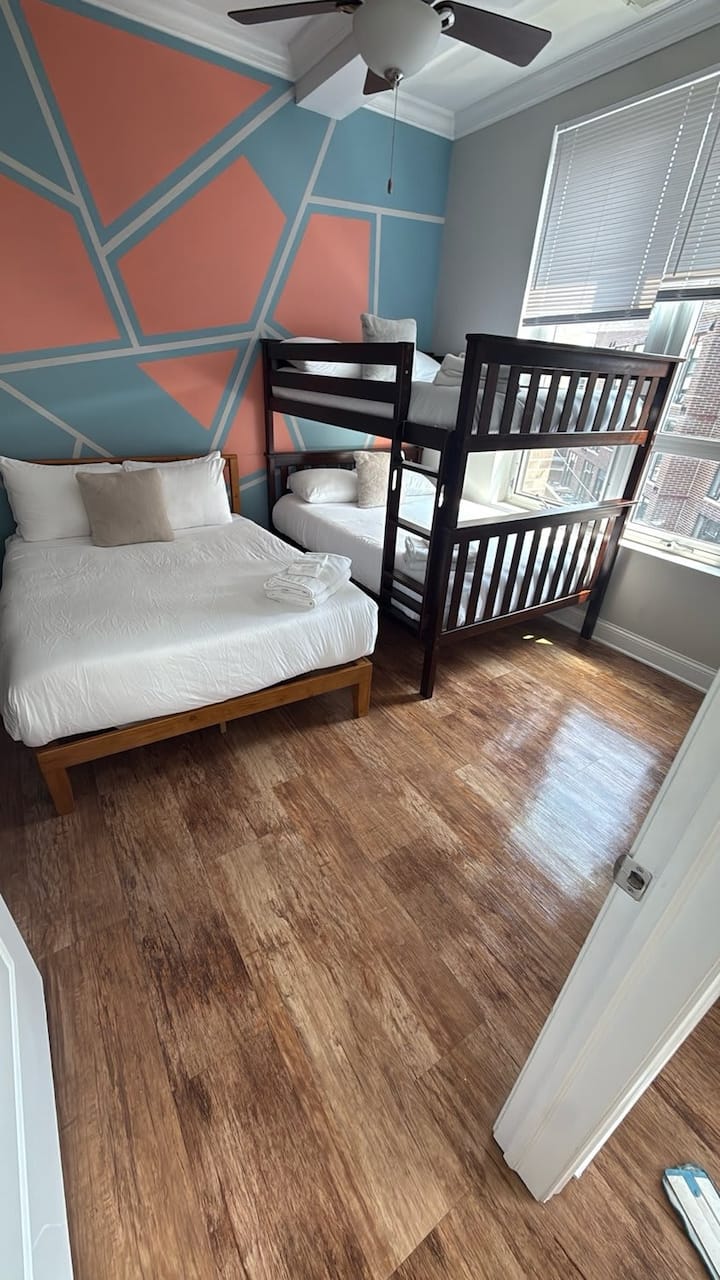 ห้องนอนที่มีชีวิตชีวาพร้อมเตียงคู่และเตียงสองชั้นติดกับผนังที่มีลวดลายเรขาคณิต ทำให้เป็นที่พักผ่อนที่มีสไตล์และอบอุ่น