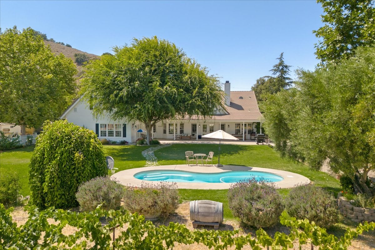 The expansive backyard features a kidney-shaped heated pool surrounded by lounge chairs. Mature trees provide shade and privacy, complementing the lush green lawn. The home's inviting exterior is framed by landscaped areas and views of the surrounding hills.