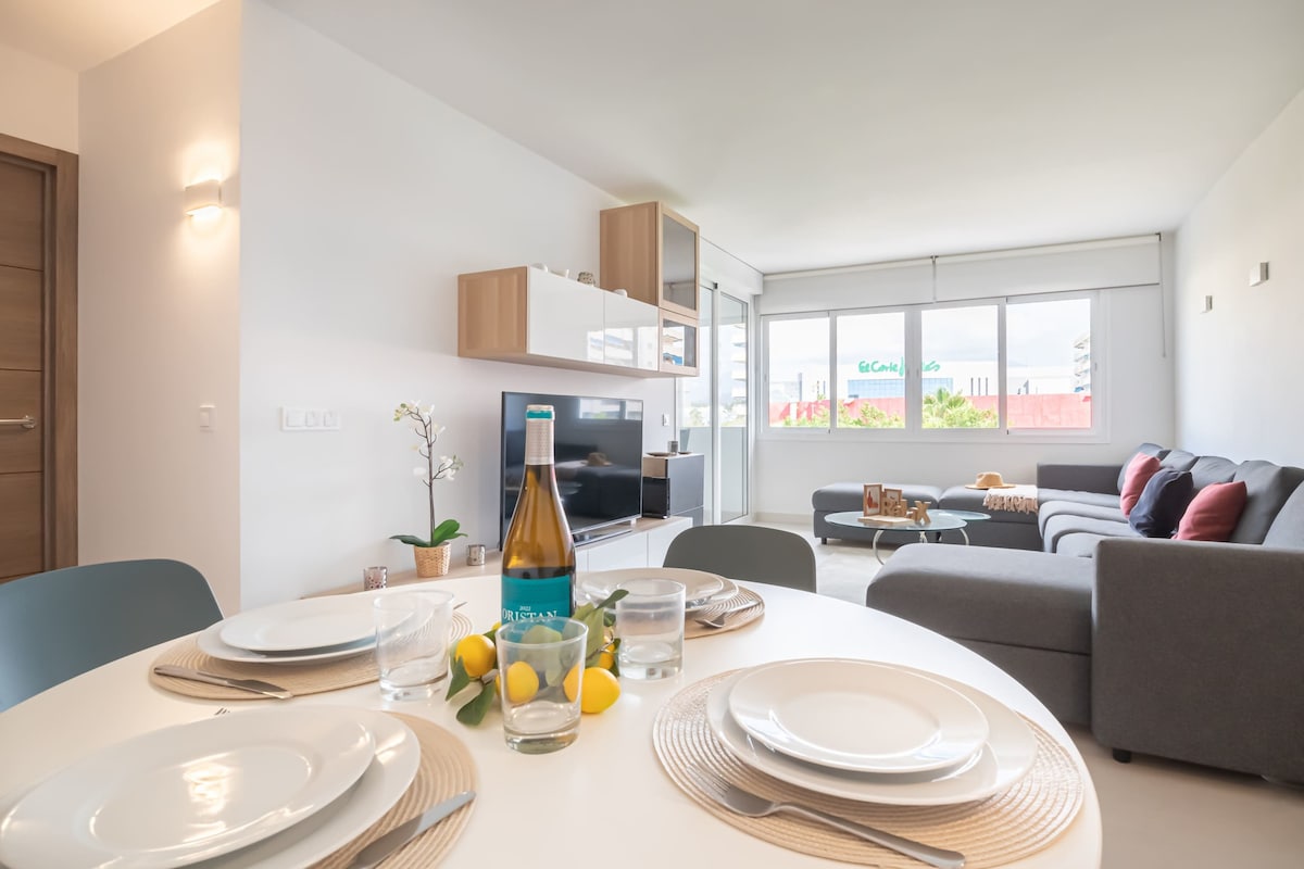 A modern dining area features a round table set with plates, glasses, and a centerpiece of lemons. In the background, a comfortable living space is visible with a gray sofa adorned with cushions and a large window allowing natural light to fill the room.