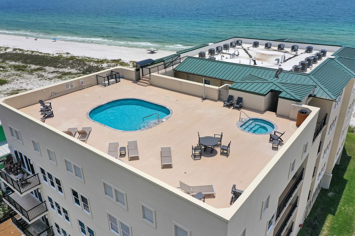 A spacious rooftop amenities area features a swimming pool and hot tub, surrounded by lounge chairs and tables. The pool deck overlooks the beach and clear waters of the Gulf, providing a relaxing space for guests to unwind and enjoy the views.