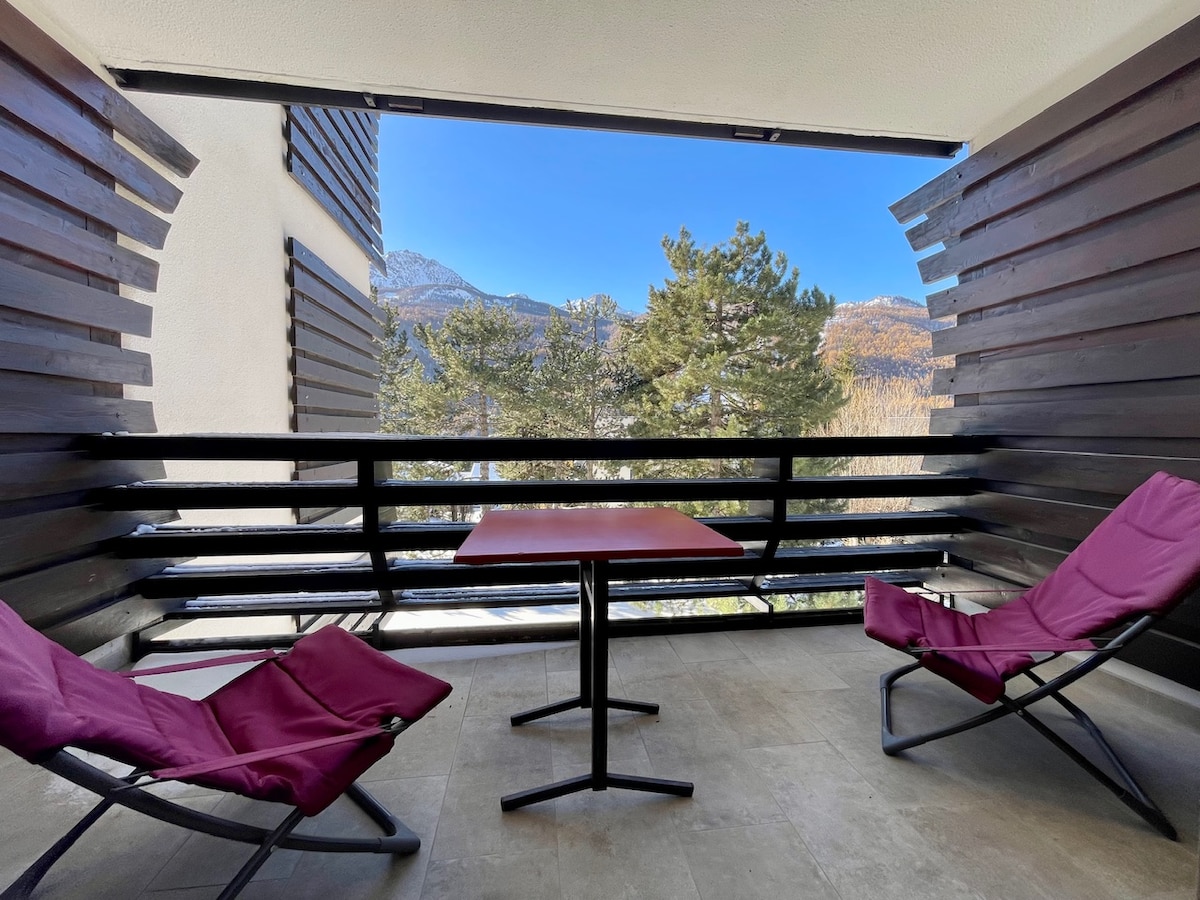 A south-facing terrace features a small red table surrounded by two maroon chairs. Wooden slats provide privacy while allowing natural light and views of the surrounding mountains and trees. The serene outdoor space invites relaxation in a tranquil setting.